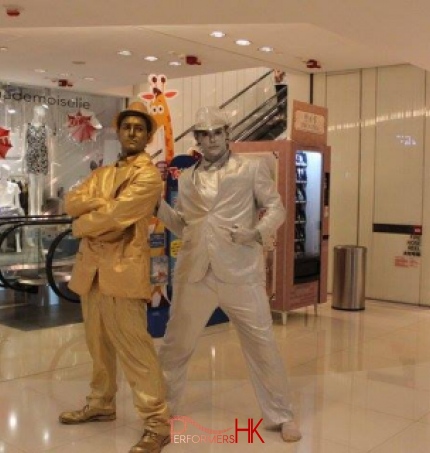 Golden statue with silver statue standing together in front of an escalator at an event 