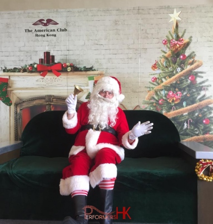 Santa in red costume sitting on black couch with backdrop of Christmas tree for American Club central, Hong Kong